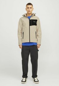 Beige jacket with a black chest pocket, two side zippers, adjustable hood, and blue accents. Paired with black pants and sneakers.