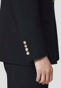 Rękaw czarnej marynarki z czterema złotymi guzikami o teksturowanym środku. Materiał jest gładki, a krój dopasowuje się do ramienia.