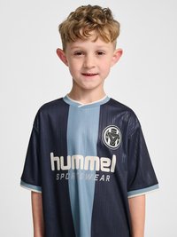 Jonge jongen met krullend haar die een donkerblauw en lichtblauw gestreept sportshirt draagt met het "hummel"-logo op de voorkant tegen een effen achtergrond.