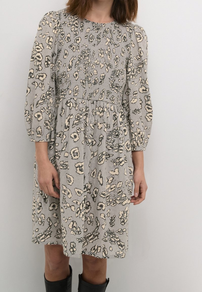 Femme portant une robe grise mi-longue avec un corsage froncé et un motif floral abstrait, associée à des bottes noires remontant jusqu'aux genoux.