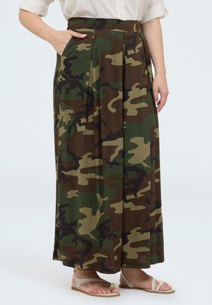 Femme portant une longue jupe camouflage avec poches latérales, chemise blanche et sandales beiges, debout avec une main dans une poche.