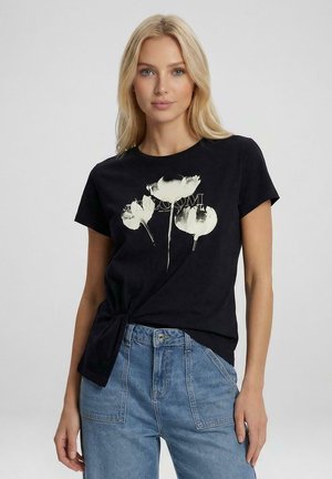 Blonde vrouw die een zwart T-shirt met witte bloemenprint en tekst "BLOOM" draagt, gecombineerd met blauw hoogwaterjeans, staand tegen een lichte achtergrond.