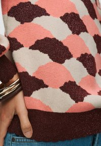 Pogled z bližine na osebo, ki nosi večbarvni pleteni pulover z roza, bež in rjavimi valovitimi vzorci ter modrimi kavbojkami, okrašenimi z zlatimi zapestnicami.