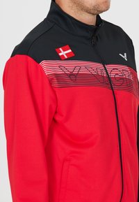 Rød og sort sportsjakke med høj krave, lynlås lukning, vandrette striber og et dansk flag patch på skulderen.