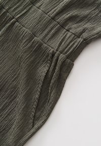 Olive green textured fabric featuring a wrinkled design with elastic detailing and a side pocket. Smooth surface with subtle sheen.