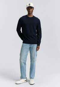 Een jonge man staat zelfverzekerd in een marineblauwe trui, lichtblauwe jeans en witte sneakers, met een crèmekleurige pet met een letter "D".