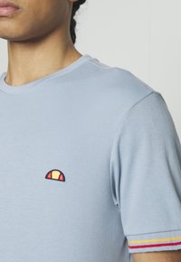 T-shirt en coton bleu clair avec un petit emblème brodé en rouge, jaune et bleu marine. Manches courtes avec des accents rayés en rouge et jaune.