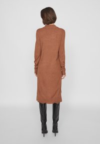 Vestito marrone a maglia con collo alto, maniche lunghe e spacchi laterali, abbinato a stivali neri al ginocchio. Tessuto liscio, lunghezza fino a metà polpaccio.