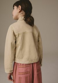 Beige farkkutakki, jossa on istuva malli, sivutaskut ja napitettavat hihansuut, yhdistetty pinkkiin raidalliseen hameeseen, jossa on läppätaskut.