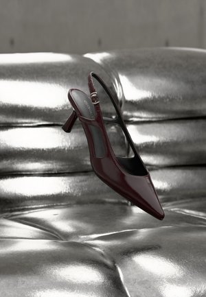 Λουστραρισμένο σκούρο κόκκινο γόβα με λουρί και ψηλό τακούνι, μυτερή μύτη, εμφανίζεται πάνω σε γυαλιστερή ασημένια επιφάνεια από δερμάτινη ταφτ.