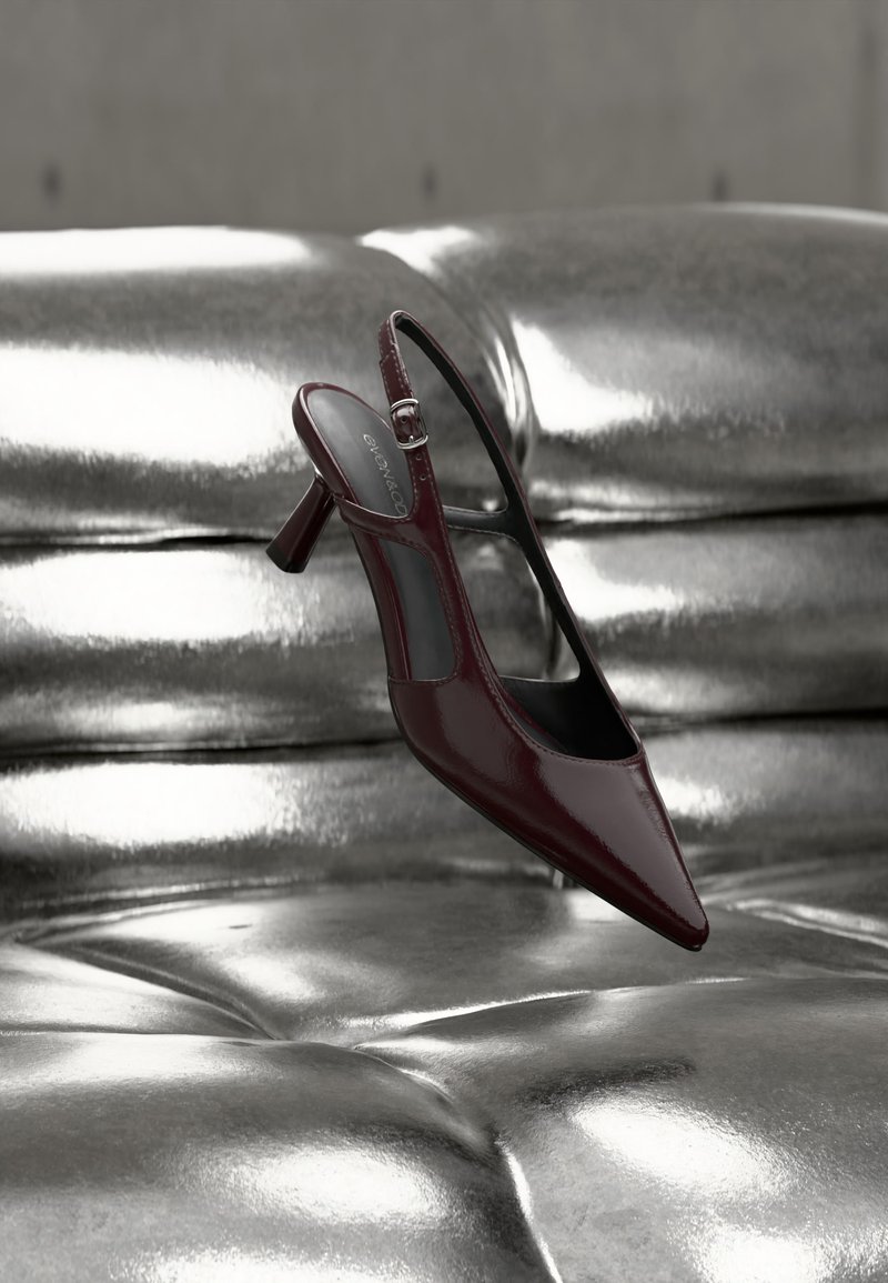 Λουστραρισμένο σκούρο κόκκινο γόβα με λουρί και ψηλό τακούνι, μυτερή μύτη, εμφανίζεται πάνω σε γυαλιστερή ασημένια επιφάνεια από δερμάτινη ταφτ.