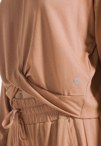 Jasnobrązowa, miękka tkanina bluzka o luźnym kroju, z przekręconym wzorem w talii oraz elastycznym pasem z sznurkiem.