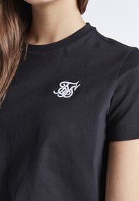 Černé bavlněné tričko s kulatým výstřihem. Má bílé vyšívané logo na levé straně hrudi. Krátké rukávy a měkký povrch.