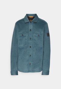 Chemise-veston bleu sarcelle à boutons, avec deux poches avant à boutons et une petite écusson noir sur la manche gauche.
