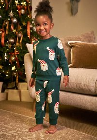 Jong kind in een feestelijke groene pyjama met kerstman gezichten staat op blote voeten in een versierde woonkamer dichtbij een kerstboom.