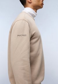 Felpa beige con collo tondo e maniche lunghe. Presenta una stampa del logo sulla manica sinistra. Realizzata in tessuto morbido e texture.