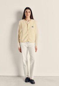 Kremo spalvos meškiuko liemenė su apvaliu kaklu, dviem priekinėmis kišenėmis ir logotipo skambe. Derinama su baltais siauro kirpimo džinsais ir juodais batais.