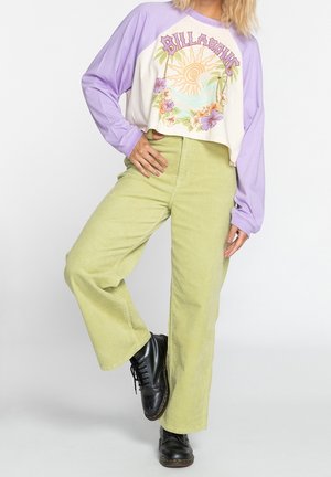 Femme portant un pantalon large vert clair, des bottes noires et une chemise à manches longues blanche et lavande avec un motif floral et solaire.
