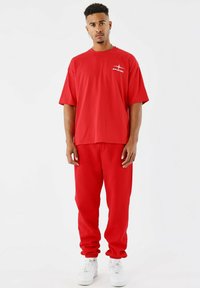 Man met kort krullend haar draagt een oversize rode t-shirt en bijpassende joggers met witte sneakers, staand tegen een witte achtergrond.