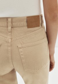 Beige spijkerbroek heeft een leren label bij de taille met gegraveerde tekst, twee achterzakken en zichtbare stikseldetails.