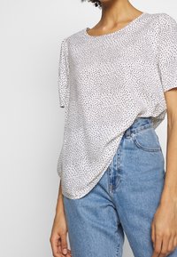 Mujer vestida con una blusa blanca de manga corta con pequeños puntos negros, metida en unos pantalones vaqueros de tiro alto de color azul claro, con la mano relajada a un lado.