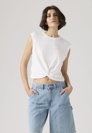 Femme aux cheveux courts bruns portant un haut blanc sans manches à devant torsadé et un short en jean bleu clair taille haute, les mains dans les poches.