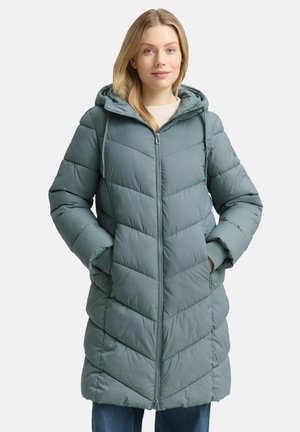 Frau trägt eine knieumspielende, türkisfarbene Steppjacke mit Kapuze, vorderem Reißverschluss und hat die Hände in den Seitentaschen, steht vor einem einfarbigen Hintergrund.