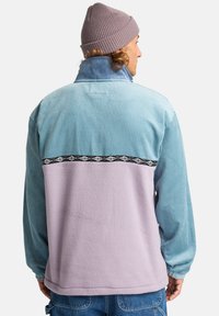 Pullover in pile e corduroy blu e lavanda, con una fascia orizzontale nera decorata sul retro. Tessuto testurizzato, colletto alto.