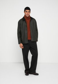 Dark green jacket with button closure, black collar, worn over a brown turtleneck. Black pants and burgundy shoes complete the outfit.