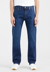 Personne portant un jean droit bleu foncé, des baskets blanches et une chemise blanche, debout devant un fond clair uni.