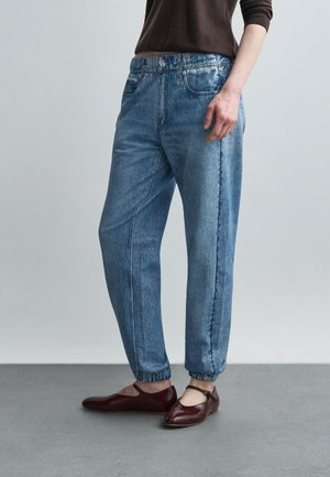 Person wearing relaxed fit blue denim jogger jeans with elastic cuffs paired with dark red Mary Jane shoes and a dark brown top.