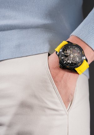 Riešas su juodu ir geltonu mechaniniu laikrodžiu, dėvimas ant šviesiai smėlio spalvos kelnių kišenės, derinamas su šviesiai mėlynu riboto megztinio rankogaliu.