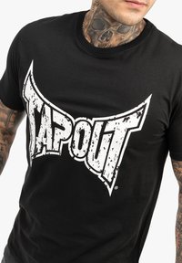 Melnas kokvilnas t-krekls ar lielu, nolietotu baltu "TAPOUT" grafiku krūšu rajonā. Īsas piedurknes, apaļa kakla izgriezums, standarta piegriezums.