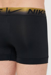 Fekete sport boxer nadrág sima textúrával és arany színátmenetes márkás derekú részsel, amelyen fekete betűkkel szerepel a "NIKE" felirat.