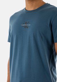 Camiseta de algodón azul con cuello redondo, que presenta un logotipo bordado en negro "Calvin Klein Jeans" y un diseño minimalista.