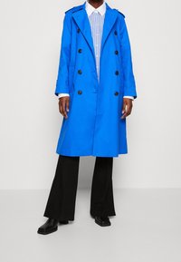 Niebieski trencz z podwójnym zapięciem na guziki i czarnymi guzikami, noszony na jasnoniebieskiej koszuli w paski i czarnych spodniach z szerokimi nogawkami.