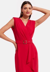 Czerwona sukienka bez rękawów z dekoltem w serek i paskiem w talii z złotą klamrą. Miękka tkanina z fasonem typu kopertowego i marszczeniami.