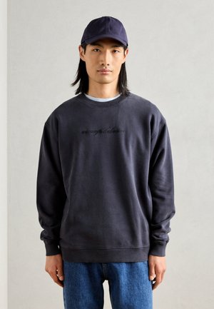 Mörkgrå bomullströja med långa ärmar, ribbad halsringning och broderad text på framsidan, ihop med blå denimjeans och en marinblå keps.