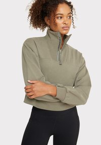 Suéter verde oliva con un cuello de media cremallera, diseño corto, puños y dobladillo acanalados, combinado con leggins negros. Textura de tela suave.