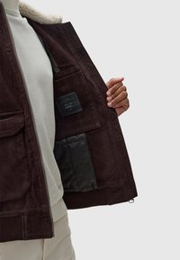 Jachetă din corduroy maro cu guler pufos, închidere cu fermoar și un buzunar interior cu căptușeală modelată.