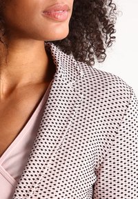 Blazer rose clair avec un motif à pois noir. Il dispose d'un col structuré et d'un tissu texturé, présenté sur un modèle partiellement visible.