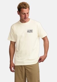 Cream cotton t-shirt featuring a rectangular graphic logo on the left chest, short sleeves, and a relaxed fit.