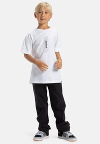 Biely bavlnený tričko s centrálnym čiernym grafickým dizajnom, spárované s voľnými čiernymi nohavicami a viacfarebnými teniskami s šedými a oranžovými akcentmi.