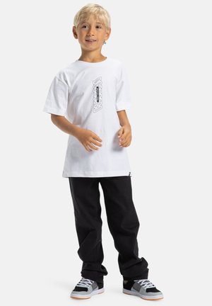 Witte katoenen t-shirt met een centraal zwart grafisch ontwerp, gecombineerd met losse zwarte broeken en meerkleurige sneakers met grijze en oranje accenten.