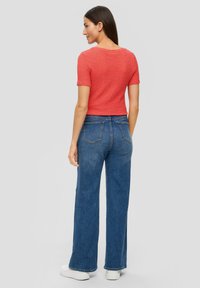 Rotes, strukturiertes T-Shirt mit kurzen Ärmeln, kombiniert mit hoch taillierten, blauen weit geschnittenen Jeans. Weiße Plateau-Sneaker sind sichtbar, der Hintergrund ist neutral grau.