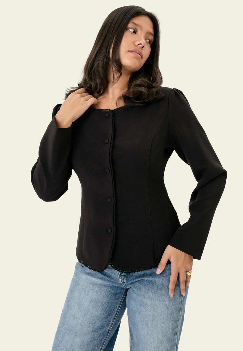 Femme aux longs cheveux foncés portant une veste noire cintrée à boutons et un jean bleu clair, posant devant un fond clair uni.