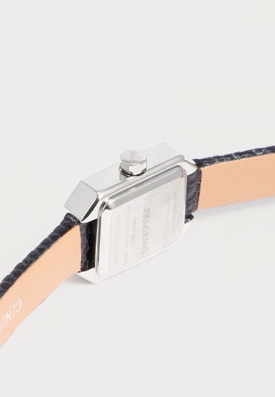 Boîtier de montre carré en argent avec couronne et bracelet en cuir noir montrant le dessous beige et les coutures.