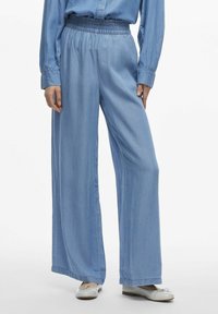 Pantaloni larghi di colore azzurro chiaro realizzati in tessuto morbido con vita elastica. Presentano una texture liscia e un orlo pulito. Abbinati a scarpe flat.