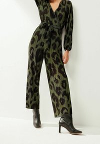 Jumpsuit verde con plisados y estampado de leopardo negro, escote en V profundo, mangas largas abullonadas y cintura atada. Combinado con botas de tobillo de tacón negro.