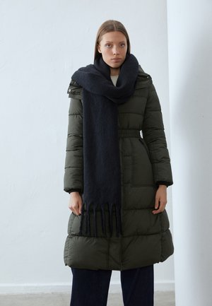 Femme portant un long manteau matelassé vert olive et une écharpe noire surdimensionnée, debout devant un mur clair uni.